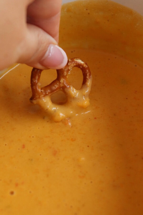 A pretzel being dipped in creamy cheese sauce in a small white crockpot.