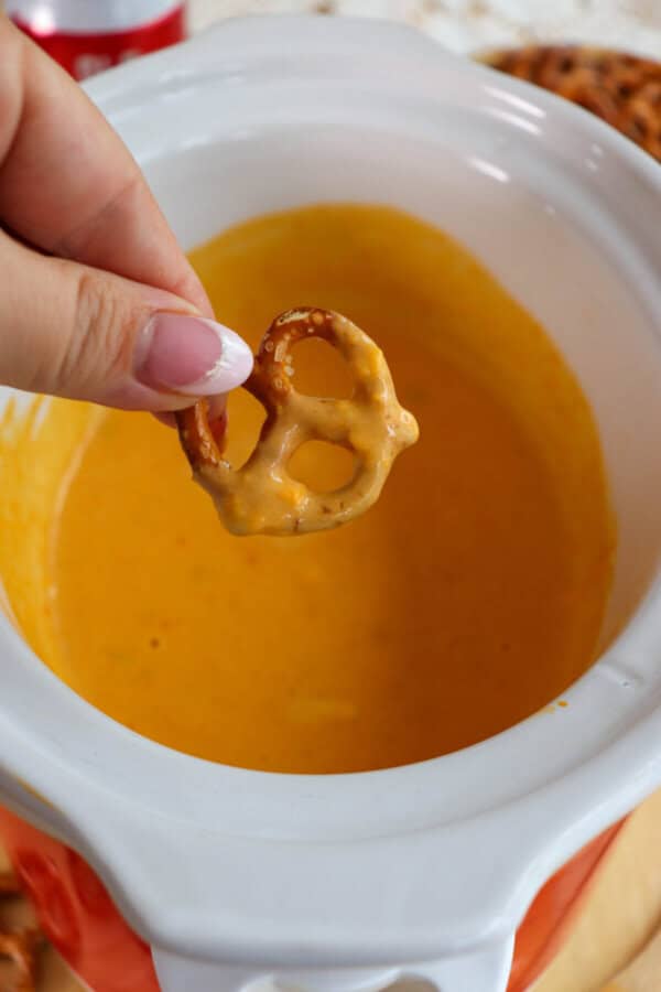 A pretzel being dipped in creamy cheese sauce in a small white crockpot.