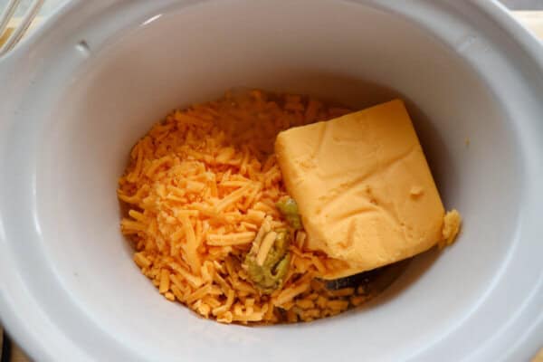 Shredded cheddar cheese, beer and a block of Velveeta cheese in a small white crockpot.