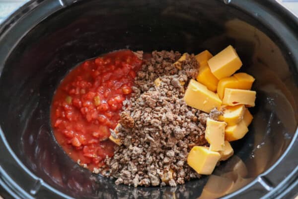 Diced tomatoes, ground beef and cubes of Velveeta in a black crockpot.