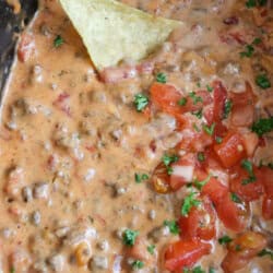 Cheesy rotel dip with diced tomatoes and ground beef in a black crockpot with a tortilla dip resting inside.