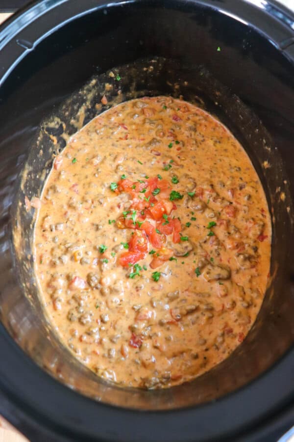 Cheesy dip with diced tomatoes and ground beef in a black crockpot topped with fresh diced tomatoes and parsley.