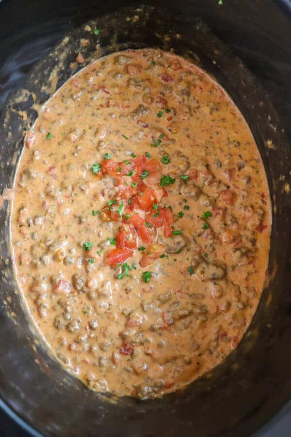 Cheesy dip with diced tomatoes and ground beef in a black crockpot topped with fresh diced tomatoes and parsley.