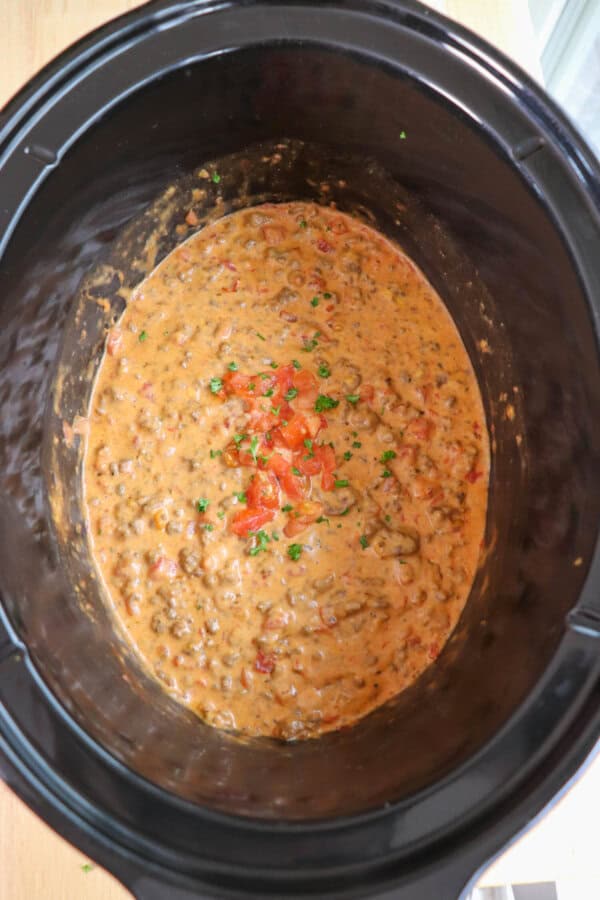 Cheesy dip with diced tomatoes and ground beef in a black crockpot topped with fresh diced tomatoes and parsley.