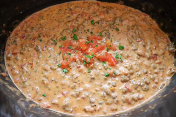 Cheesy dip with diced tomatoes and ground beef in a black crockpot topped with fresh diced tomatoes and parsley.