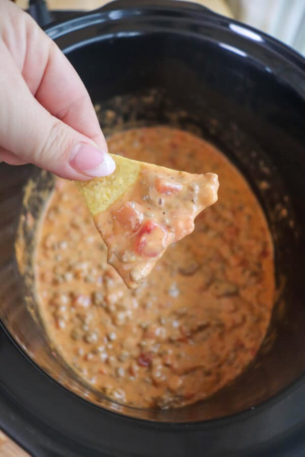 Cheesy dip with diced tomatoes and ground beef in a black crockpot topped with fresh diced tomatoes and parsley with a tortilla being dipped in and held by a hand.