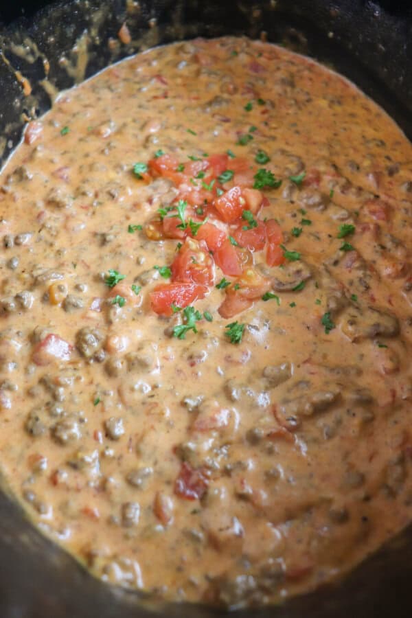 Cheesy dip with diced tomatoes and ground beef in a black crockpot topped with fresh tomatoes and parsley.