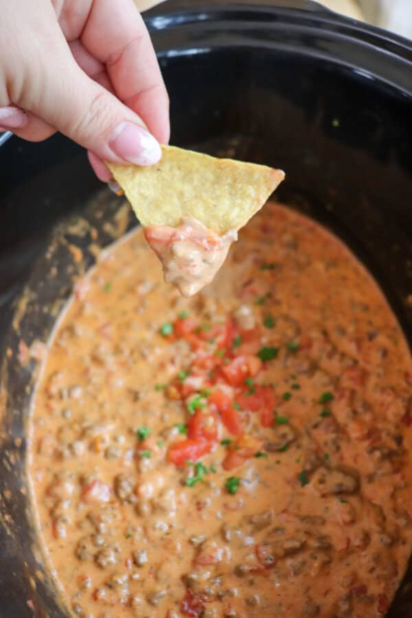 Cheesy dip with diced tomatoes and ground beef in a black crockpot topped with fresh tomatoes and parsley with a tortilla being dipped and held in a hand.