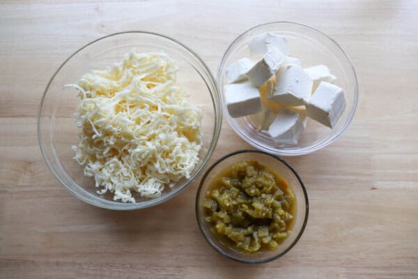 Cubed cream cheese, shredded pepper jack cheese and diced chilis in clear glass bowls on a wooden table.