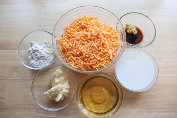 Ingredients for beer cheese sauce in clear glass bowls on a wooden table.