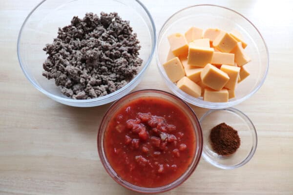 Ground beef, cubed velveeta, rotel and ground chili spice in clear glass bowls on a wooden table.