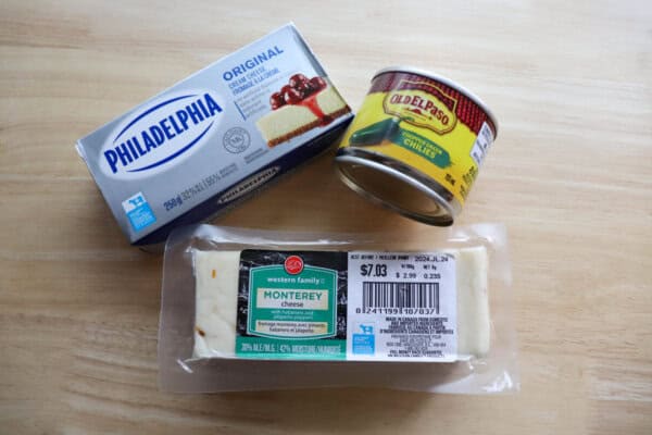 Philadelphia cream cheese in the package, old el paso diced chilis in a can and a block of white monterey jack cheese on a wooden table.