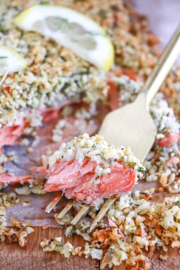Salmon fillet covered in a crust of panko and herbs on a sheet of parchment paper topped with lemon slices for garnish with a fork holding a piece.