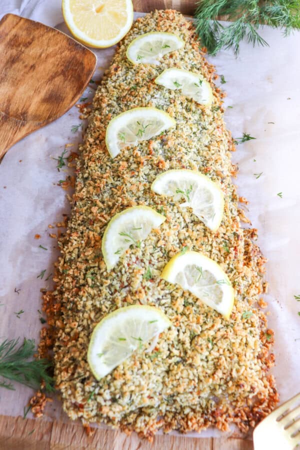 Salmon fillet covered in a crust of panko and herbs on a sheet of parchment paper topped with lemon slices for garnish.