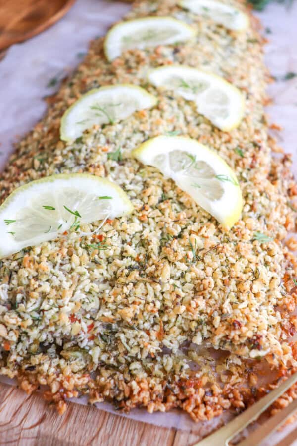 Salmon fillet covered in a crust of panko and herbs on a sheet of parchment paper topped with lemon slices for garnish.
