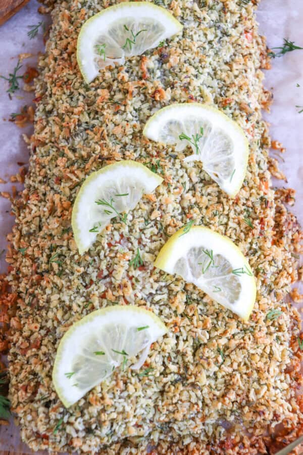 Salmon fillet covered in a crust of panko and herbs on a sheet of parchment paper topped with lemon slices for garnish.