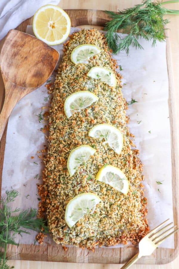 Salmon fillet covered in a crust of panko and herbs on a sheet of parchment paper topped with lemon slices for garnish.