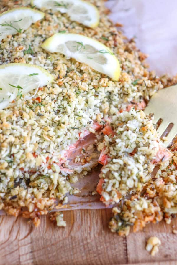 Salmon fillet covered in a crust of panko and herbs on a sheet of parchment paper topped with lemon slices for garnish with a fork in a piece.
