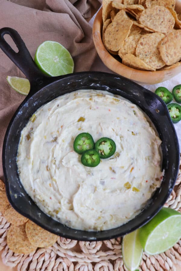 White pepper jack cheese dip in a cast iron skillet topped with three slices of jalapeno pepper and surrounded by tortilla chips and fresh lime on the table.