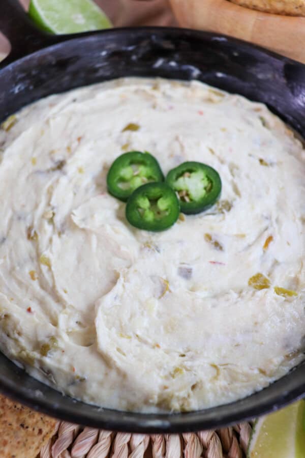 Close up of white pepper jack cheese dip in a cast iron skillet topped with three slices of jalapeno pepper and surrounded by tortilla chips and fresh lime on the table.