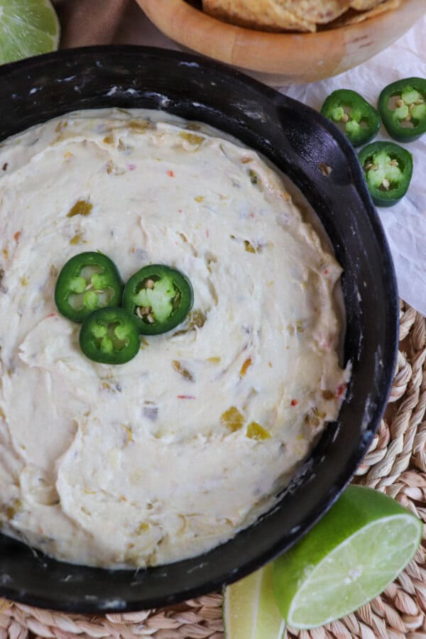 White pepper jack cheese dip in a cast iron skillet topped with three slices of jalapeno pepper and surrounded by tortilla chips and fresh lime on the table.