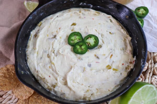 White pepper jack cheese dip in a cast iron skillet topped with three slices of jalapeno pepper and surrounded by tortilla chips and fresh lime on the table.