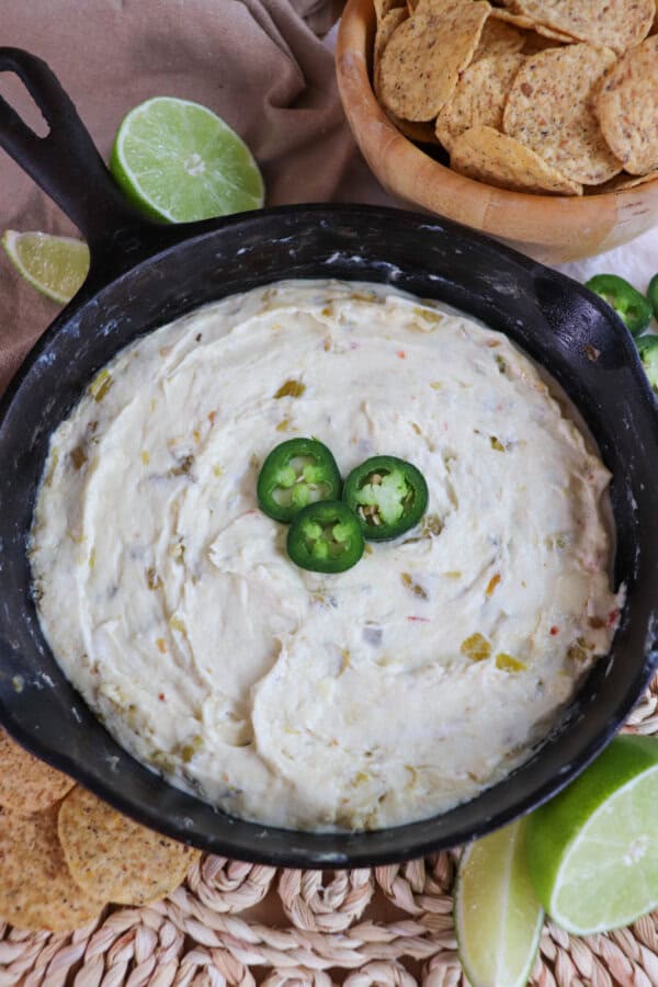 White pepper jack cheese dip in a cast iron skillet topped with three slices of jalapeno pepper and surrounded by tortilla chips and fresh lime on the table.