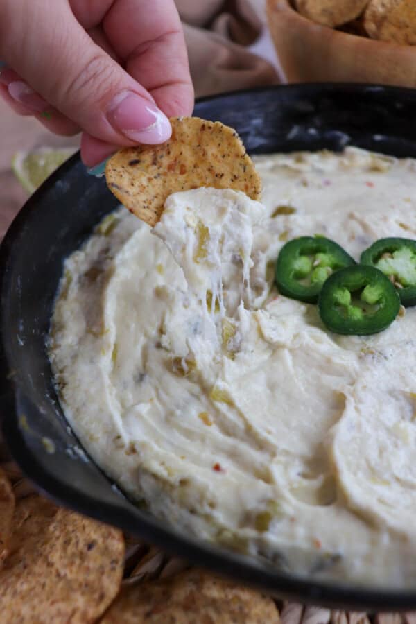 White pepper jack cheese dip in a cast iron skillet topped with three slices of jalapeno pepper with a tortilla chip pulling the cheese from the dish.