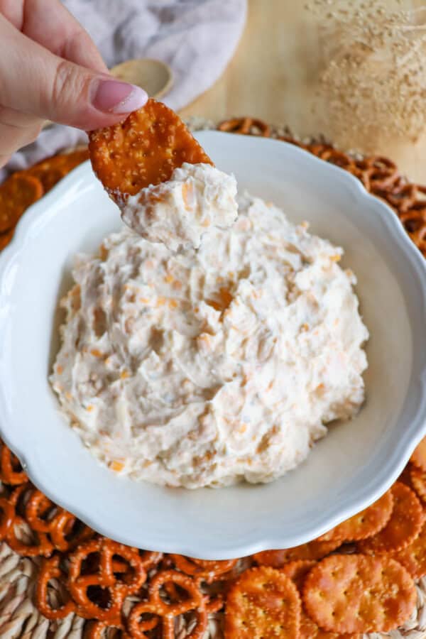 Creamy ranch beer cheese dip in a white bowl, surrounded by pretzels and crackers with a hand holding a pretzel cracker with dip on it.