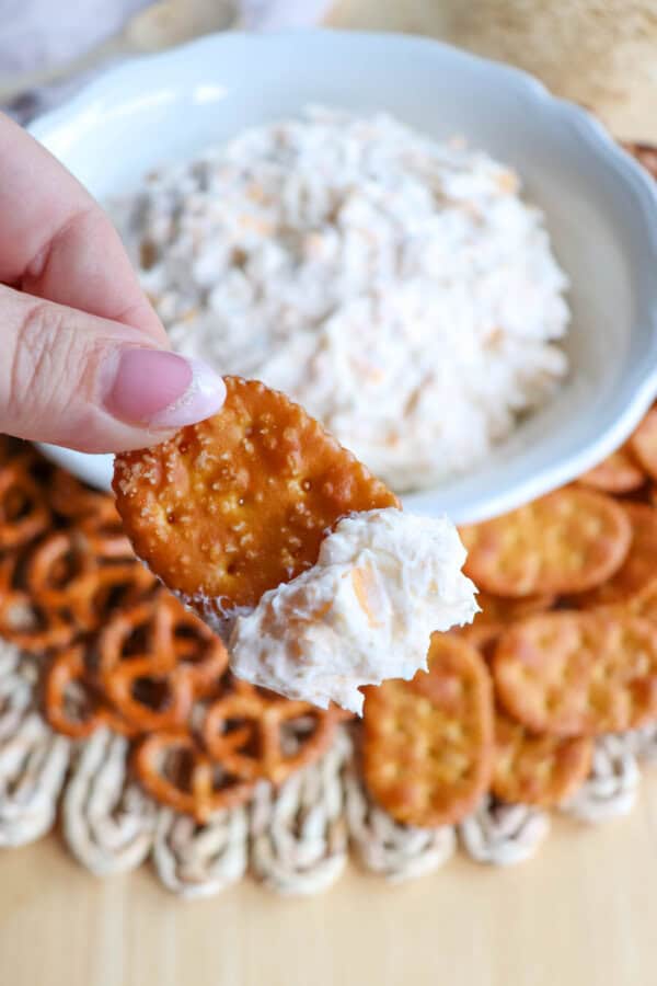 Creamy ranch beer cheese dip in a white bowl, surrounded by pretzels and crackers with a hand holding a pretzel cracker with dip on it.