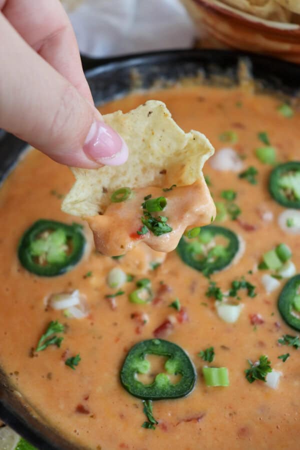 Cheesy rotel dip with no meat in a tortilla scoop held in hand with dip in the background topped with sliced jalapenos and diced green onion.
