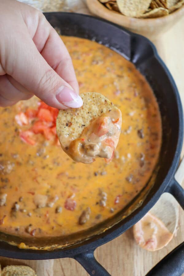 Creamy cheese dip with ground beef and tomatoes in a cast iron skillet with a round tortilla chip being dipped and held in hand.