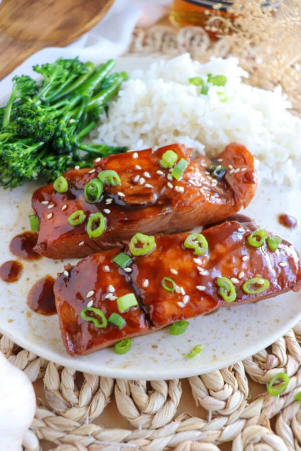 Salmon fillets covered in a brown sugar & bourbon glaze on a white plate with green onion and sesame seeds garnishing the fish.