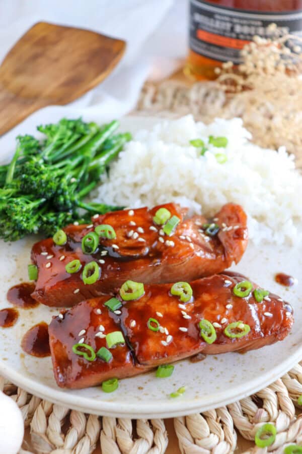 Salmon fillets covered in a brown sugar & bourbon glaze on a white plate with green onion and sesame seeds garnishing the fish.