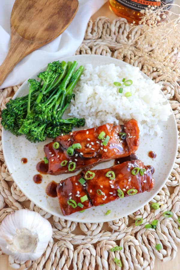 Salmon fillets covered in a brown sugar & bourbon glaze on a white plate with green onion and sesame seeds garnishing the fish.