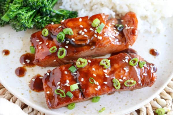 Salmon fillets covered in a brown sugar & bourbon glaze on a white plate with green onion and sesame seeds garnishing the fish.