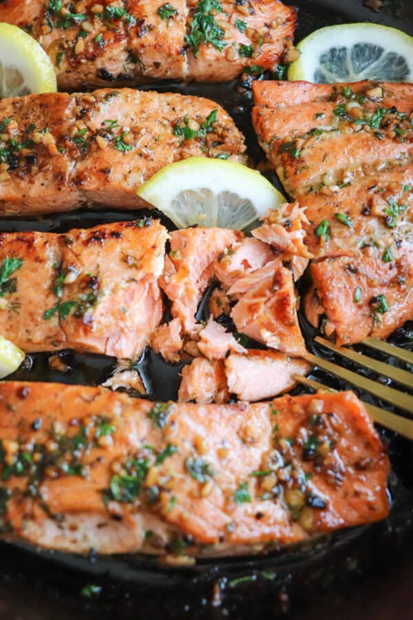 Salmon fillets in a cast iron skillet topped with fresh parsley and lemon slices and a gold fork taking a piece.