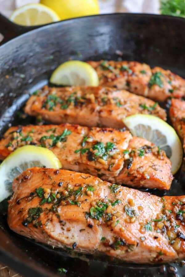 Salmon fillets in a cast iron skillet topped with fresh parsley and lemon slices.