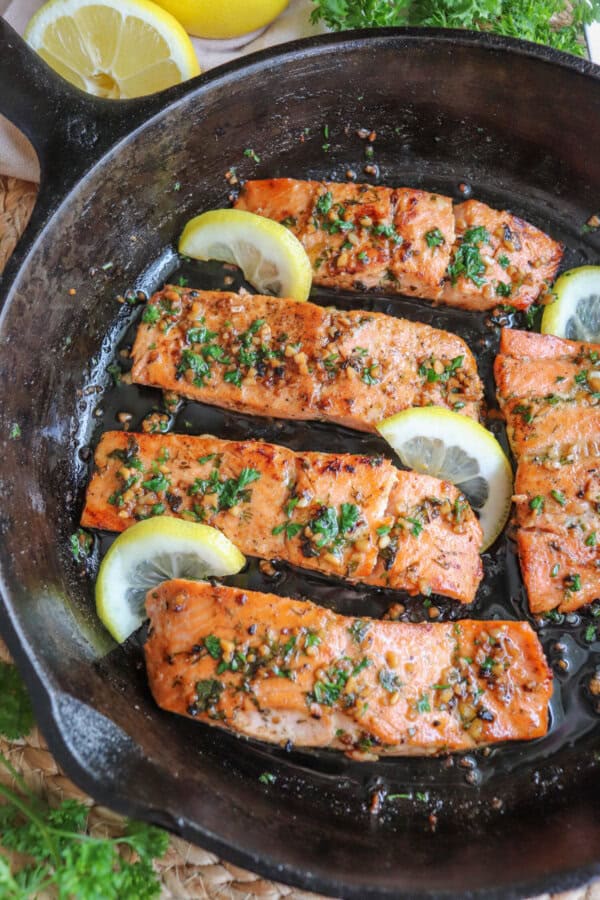 Salmon fillets in a cast iron skillet topped with fresh parsley and lemon slices.
