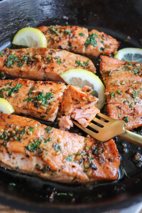 Salmon fillets in a cast iron skillet topped with fresh parsley and lemon slices with a fork in it.