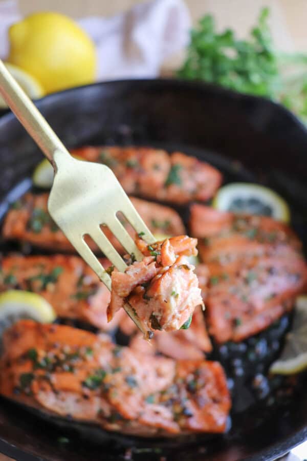 Salmon fillets in a cast iron skillet topped with fresh parsley and lemon slices and a fork full of salmon.