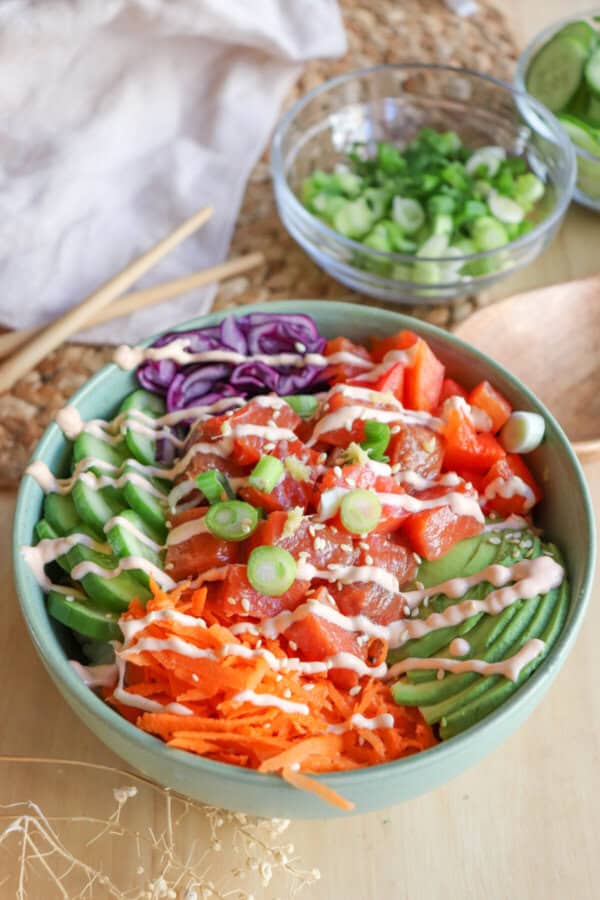 A green bowl with sliced cucumbers, grated carrot, sliced avocado, purple cabbage, diced red pepper with raw cubes of salmon topped with a drizzle of spicy mayo and diced green onion.