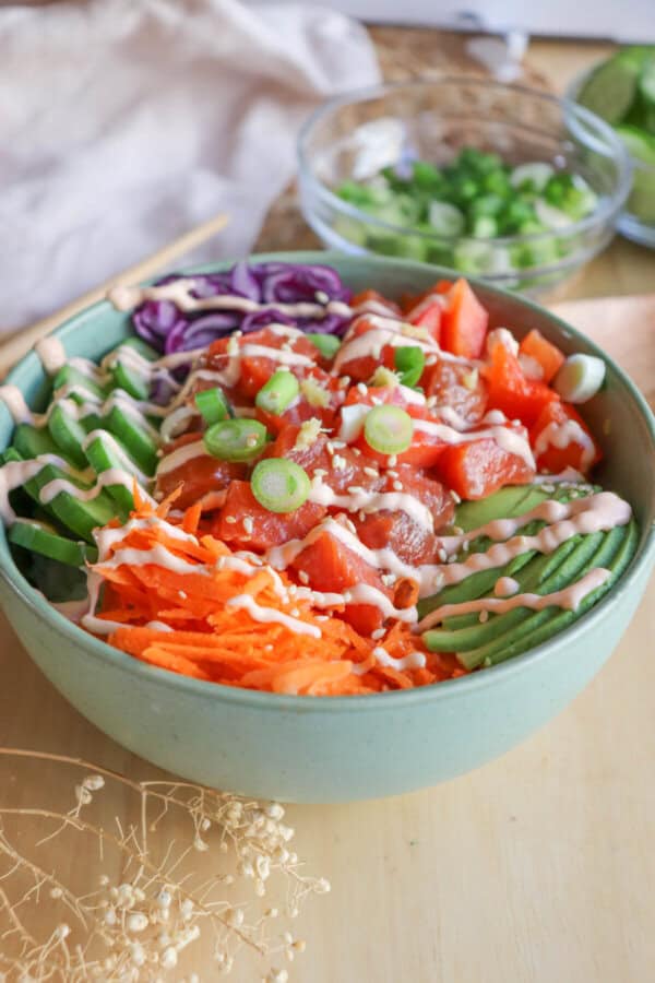 A green bowl with sliced cucumbers, grated carrot, sliced avocado, purple cabbage, diced red pepper with raw cubes of salmon topped with a drizzle of spicy mayo and diced green onion.