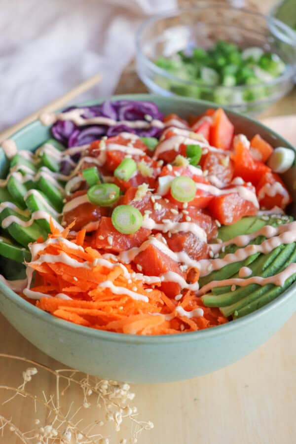 A green bowl with sliced cucumbers, grated carrot, sliced avocado, purple cabbage, diced red pepper with raw cubes of salmon topped with a drizzle of spicy mayo and diced green onion.