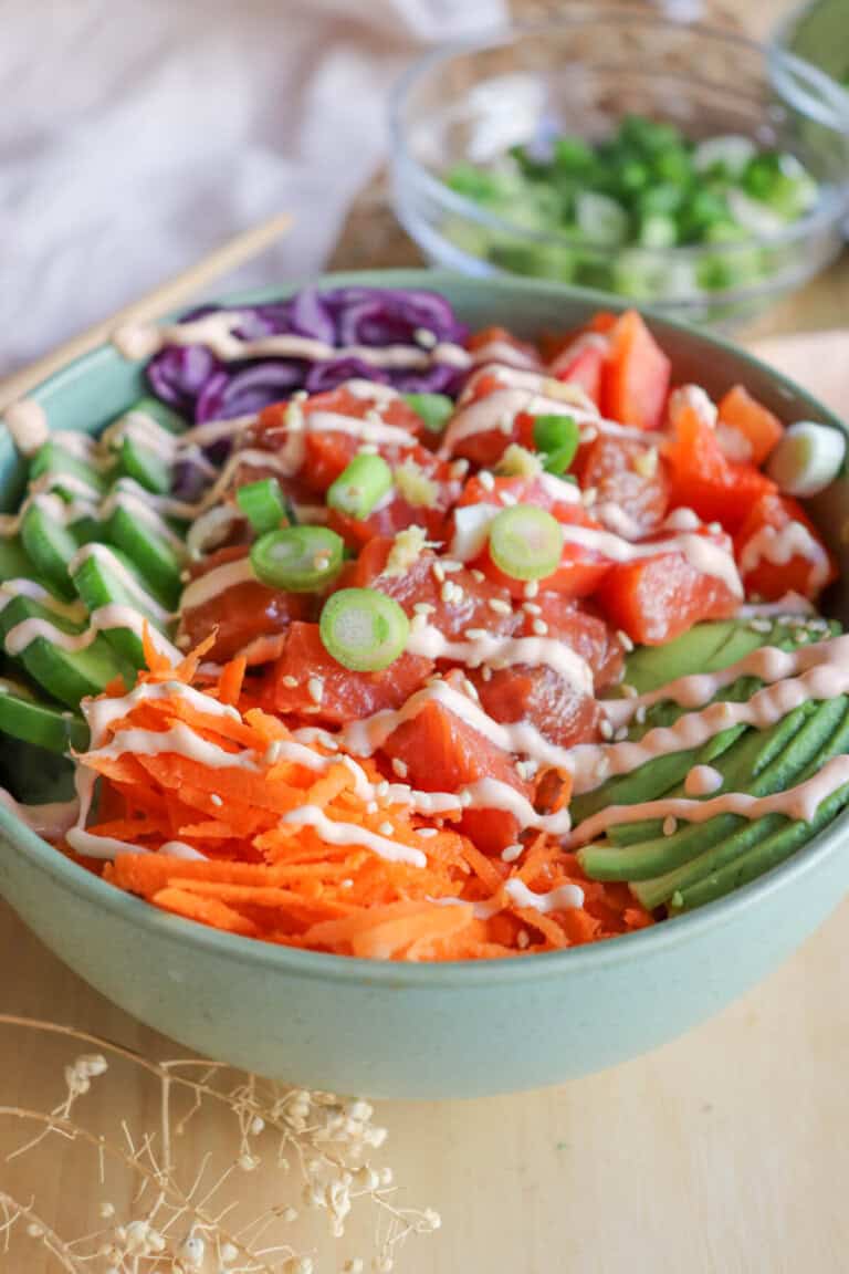 A green bowl with sliced cucumbers, grated carrot, sliced avocado, purple cabbage, diced red pepper with raw cubes of salmon topped with a drizzle of spicy mayo and diced green onion.