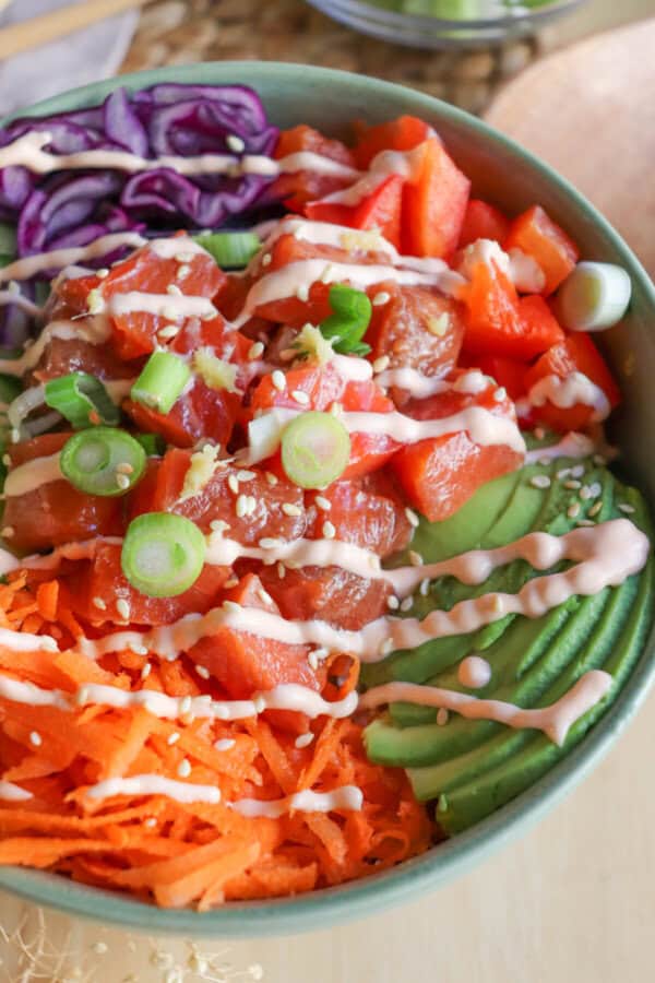 Close up of a green bowl with sliced cucumbers, grated carrot, sliced avocado, purple cabbage, diced red pepper with raw cubes of salmon topped with a drizzle of spicy mayo and diced green onion.