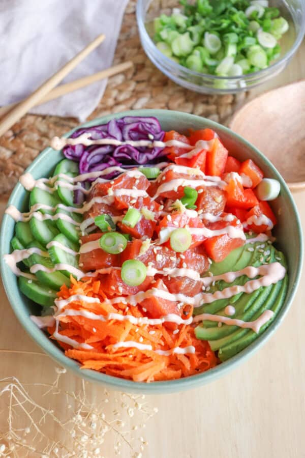 A green bowl with sliced cucumbers, grated carrot, sliced avocado, purple cabbage, diced red pepper with raw cubes of salmon topped with a drizzle of spicy mayo and diced green onion.