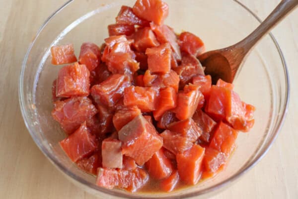 Raw salmon in marinade in a clear glass bowl with wooden spoon.
