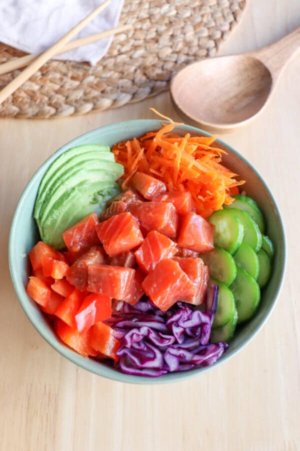 A green bowl with sliced cucumbers, grated carrot, sliced avocado, purple cabbage, diced red pepper with raw cubes of salmon.