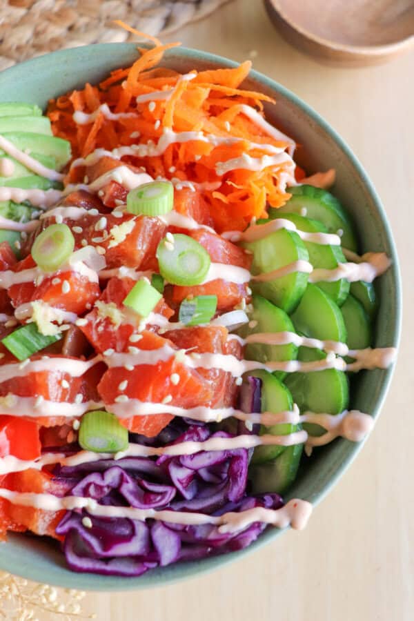Close up of a green bowl with sliced cucumbers, grated carrot, sliced avocado, purple cabbage, diced red pepper with raw cubes of salmon topped with a drizzle of spicy mayo and diced green onion.